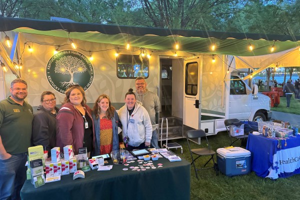A Vital Strategies team stands and smiles in front of a mobile healthcare van at a mobile harm reduction site. The van has a large awning with string lights. A table displays promotional materials and healthcare items. Trees and additional tables are visible in the background.