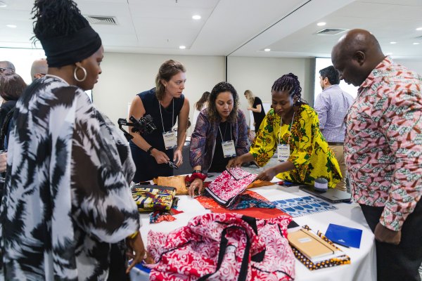 A group of people at a weaving resilience convening gathered around a table display, examining colorful patterned fabrics and handmade items at a conference or workshop in a bright, modern room.